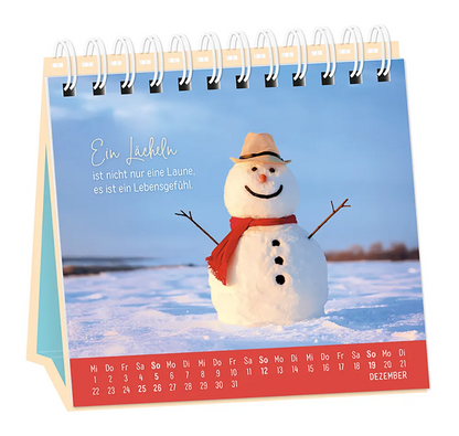 Tischkalender - "Extraportion Glück" - 2027 - Monat Dezember -  Spruch: "Ein Lächeln ist nicht nur eine Laune, es ist ein Lebensgefühl." Design: Ein lächelnder Schneemann mit Schal und Hut in einer Schneelandschaft.