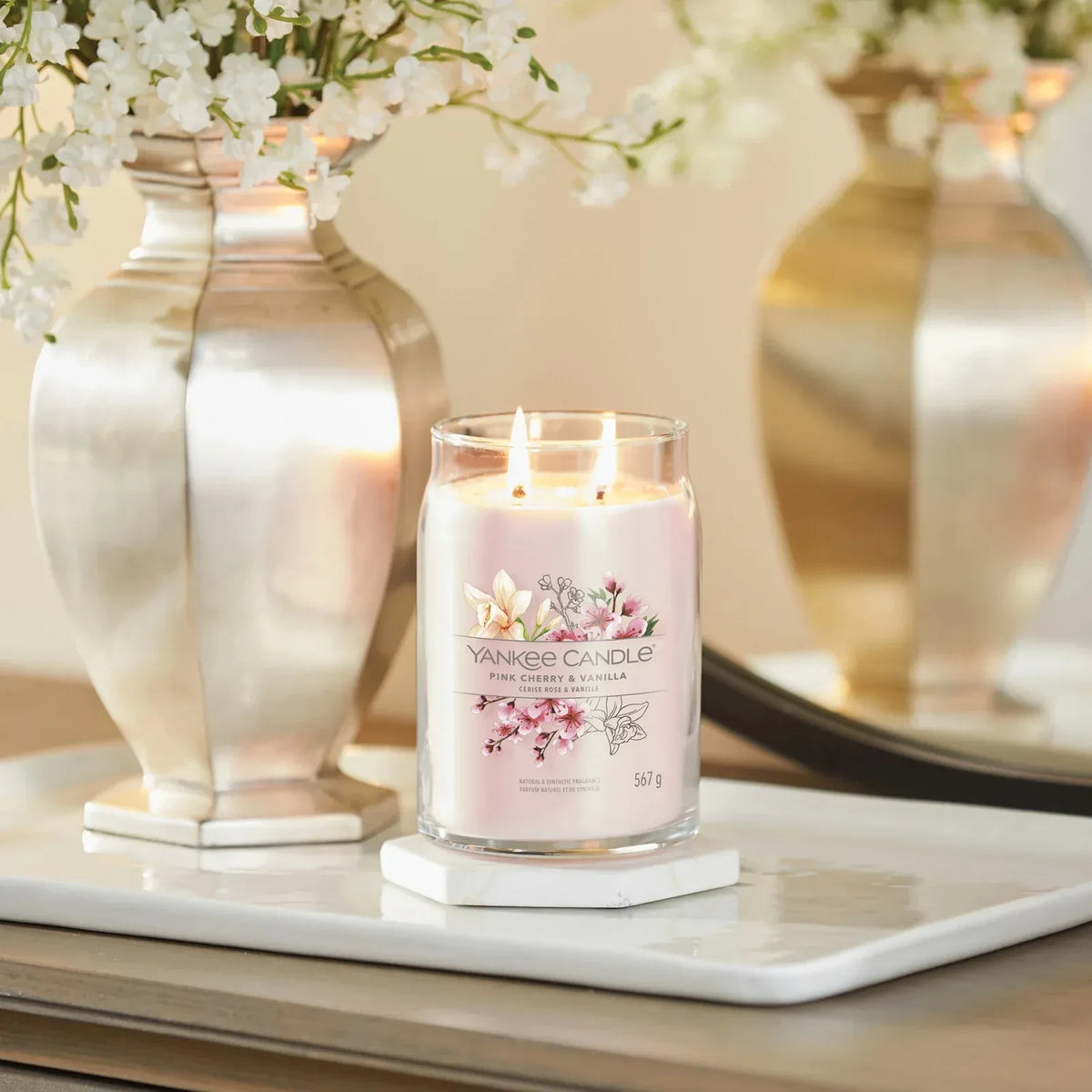 Brennende Yankee Candle Duftkerze „Pink Cherry & Vanilla“ im Glas mit zwei Dochten neben einer Vase mit weißen Blüten