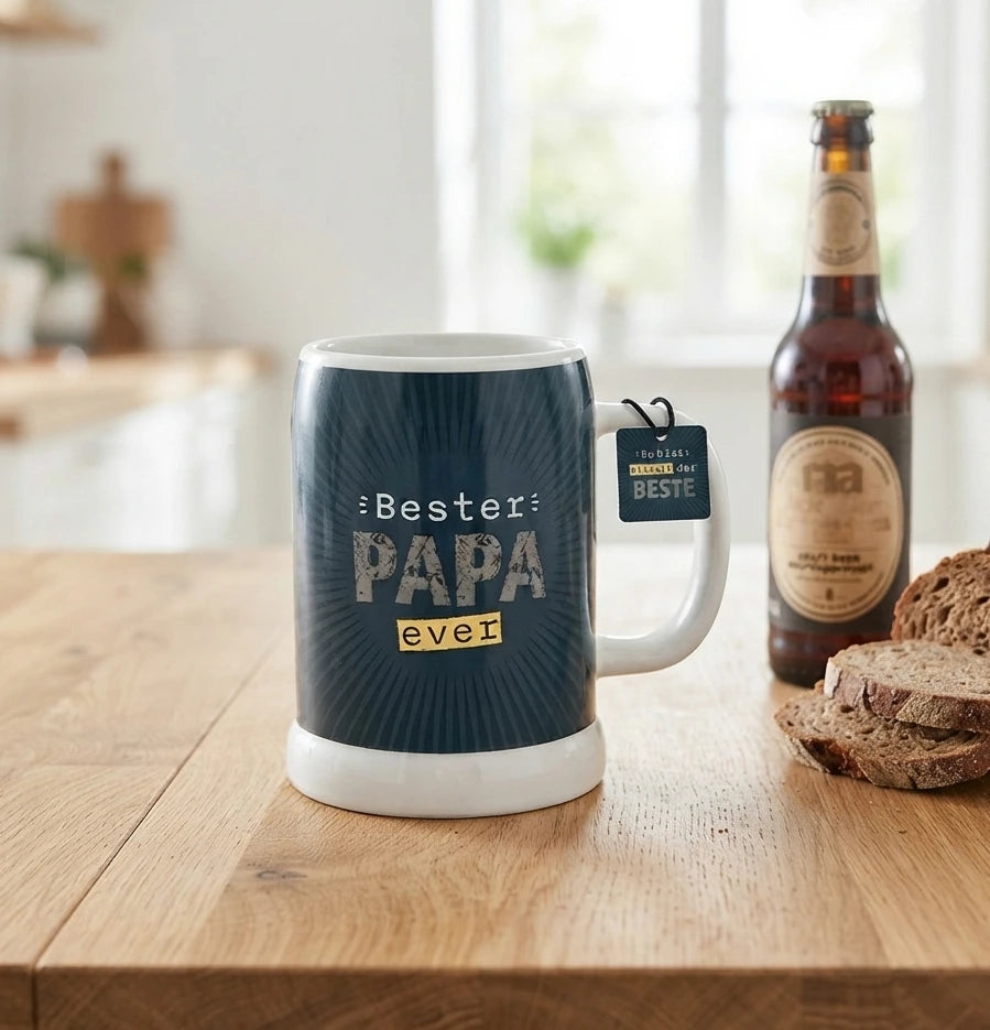 Ein blauer Bierkrug mit der Aufschrift „Bester PAPA ever“ steht neben einer Bierflasche und Brot auf einem Holztisch