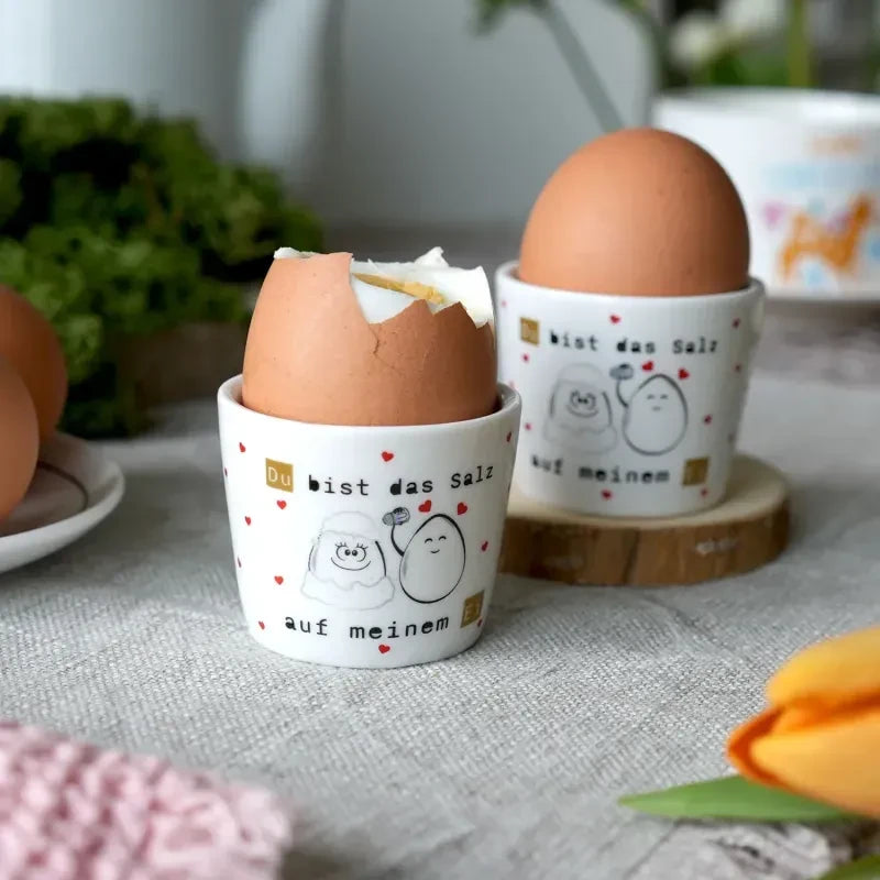 Zwei Sheepworld-Eierbecher mit Eiern, Schaf- und Eiermotiven sowie dem Spruch „Du bist das Salz auf meinem Ei“ auf einem Frühstückstisch