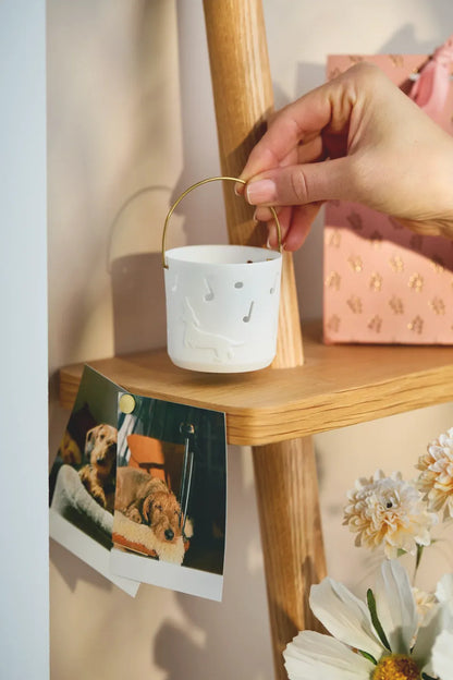 Räder Geschenklaterne „Time to be happy“ aus weißem Porzellan mit Hundemotiv und Musiknoten, am goldfarbenen Henkel über einem Holzregal gehalten
