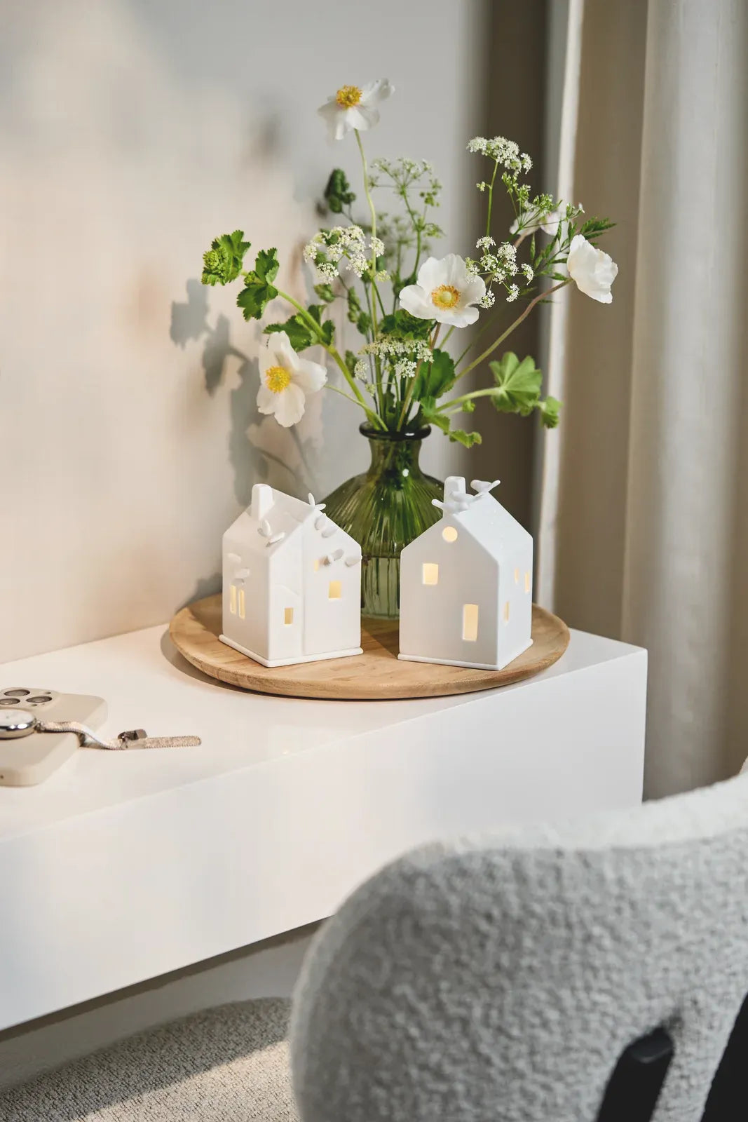 Zwei weiße räder Lichthäuser aus Porzellan auf einem runden Holztablett vor einer grünen Glasvase mit weißen Blumen
