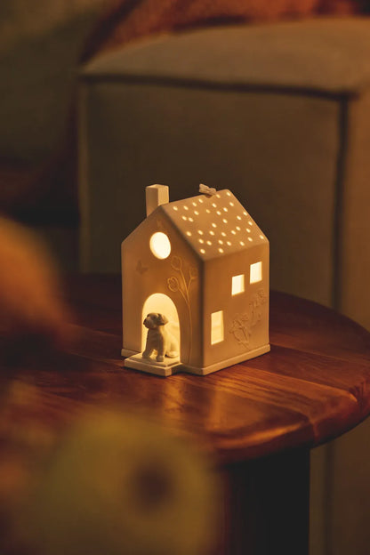 Warm beleuchtetes räder Lichthaus „Hund“ aus Porzellan mit Hundefigur im Eingang und Lichtpunkten im Dach auf einem Holztisch