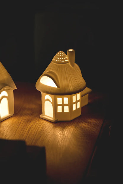 Warm beleuchtetes räder Lichthaus „Reetdachturm“ aus Porzellan mit leuchtenden Fenstern, Dachgaube und Lichtpunkten im Turmdach auf einem Holztisch
