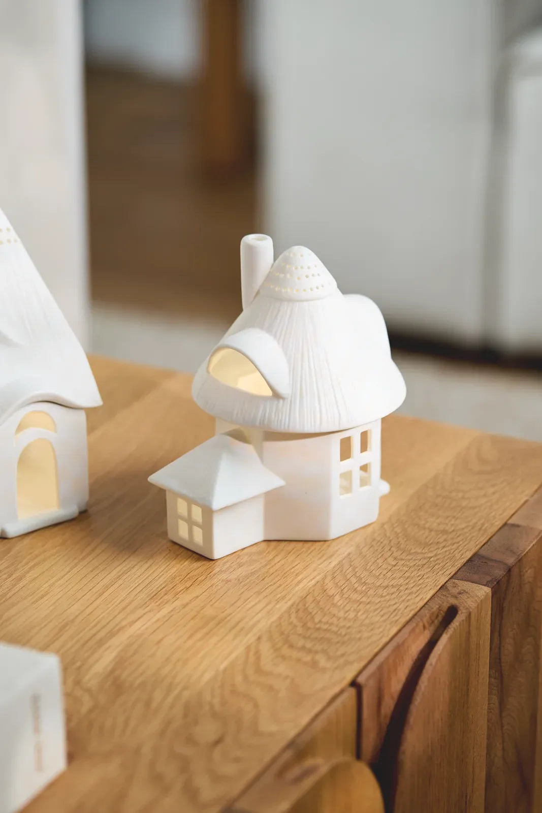 Weißes räder Lichthaus „Reetdachturm“ aus Porzellan mit kleinem Anbau und beleuchteter Dachgaube auf einer Holzkommode bei Tageslicht